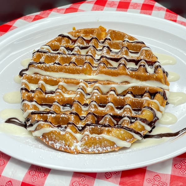 A golden waffle drizzled with white and dark chocolate sauces, dusted with powdered sugar on a white plate.