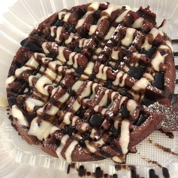 A chocolate waffle with white and dark chocolate drizzle, and powdered sugar, in a clear plastic container.