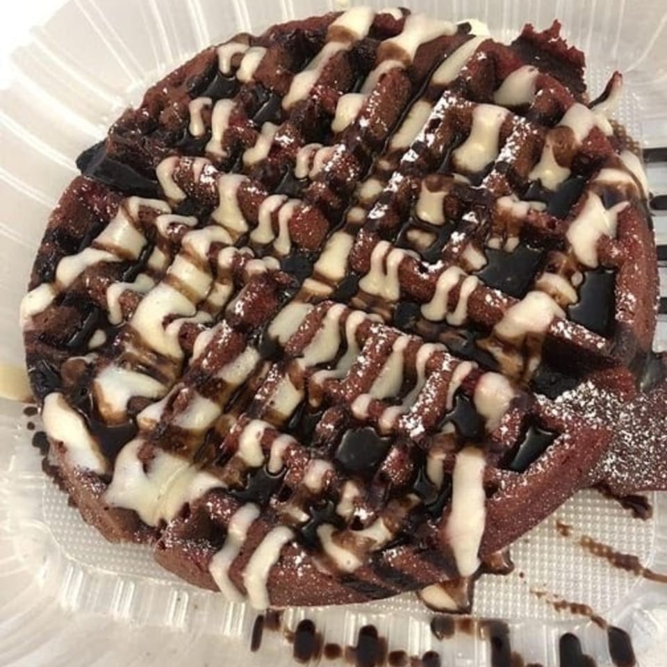 A chocolate waffle with white and dark chocolate drizzle, and powdered sugar, in a clear plastic container.